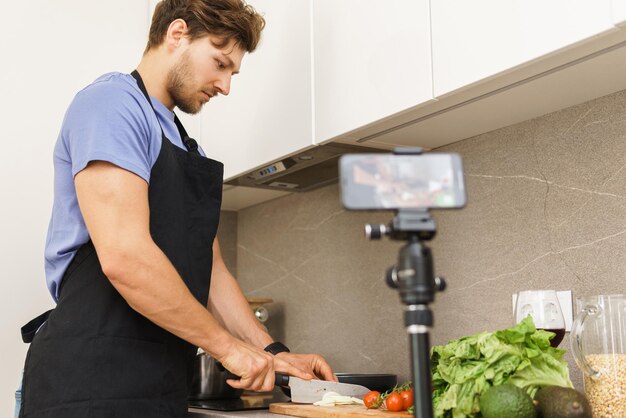 Le jeune blogueur chef barbu coupe des oignons avec un couteau sur une planche à découper dans une cuisine devant la caméra