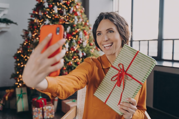 Jeune blogueuse en ligne italienne faisant selfie avec un cadeau de Noël sur fond d'arbre de Noël