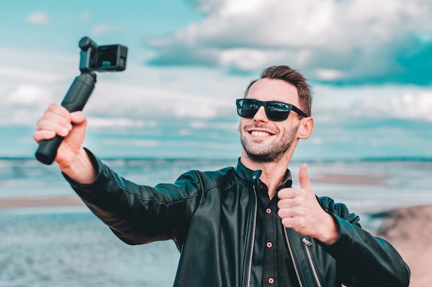 Jeune blogueuse en lunettes de soleil faisant un selfie ou une vidéo en streaming à la plage à l'aide d'une caméra d'action avec stabilisateur de caméra à cardan.