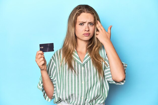 Une jeune blonde avec une carte de crédit dans un studio bleu montrant un geste de déception avec l'index