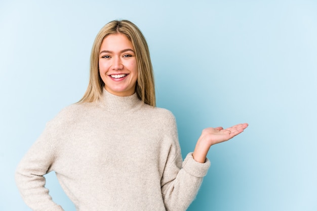 Jeune blonde caucasienne femme isolée montrant un espace de copie sur une paume et tenant une autre main sur la taille.