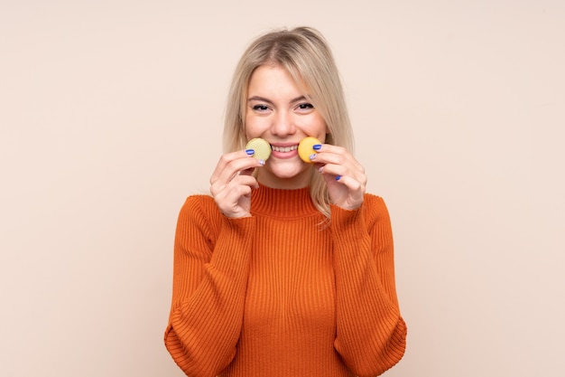 Jeune blonde femme russe sur un mur isolé tenant des macarons français colorés et souriant