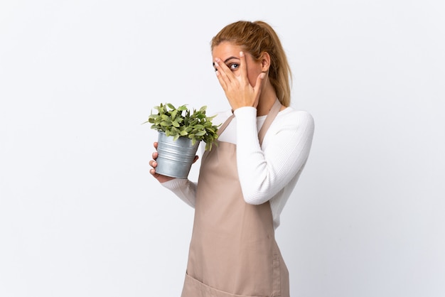 Jeune blonde jardinier femme femme tenant une plante sur un mur blanc isolé couvrant les yeux et regardant à travers les doigts