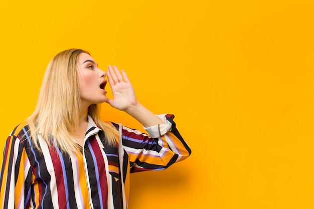 Jeune blonde jolie femme crier haut et fort pour copier l'espace sur le côté, avec la main à côté de la bouche contre le mur orange