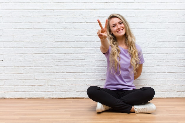 Jeune blonde jolie femme souriante et heureuse, insouciante et positive, gesticulant victoire ou paix d'une main