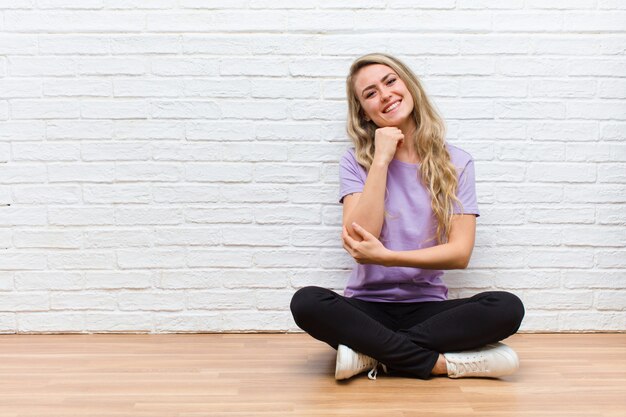 Jeune blonde jolie femme souriante, profitant de la vie, se sentant heureuse, amicale, satisfaite et insouciante avec la main sur le menton