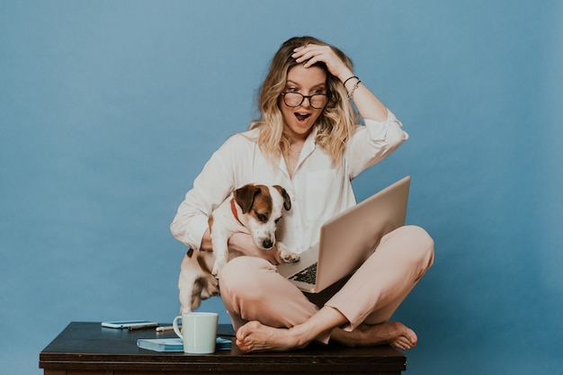 Jeune blonde à lunettes, vêtue d'une chemise blanche et d'un pantalon rose clair, assise sur une table avec un ordinateur portable, étreignant son chiot Jack Russell, elle est choquée par la nouvelle avec les yeux et la bouche grands ouverts.