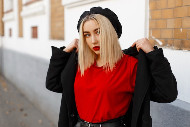 Jeune blonde moderne avec une coiffure avec des lèvres sexy rouges dans un élégant béret noir dans un long manteau à la mode dans un T-shirt rouge posant près d'un vieux bâtiment