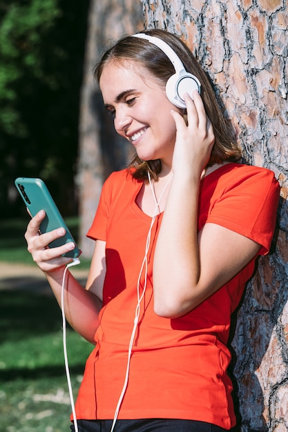 Jeune blonde portant une chemise rouge regarde le téléphone portable et écoute de la musique dans un parc