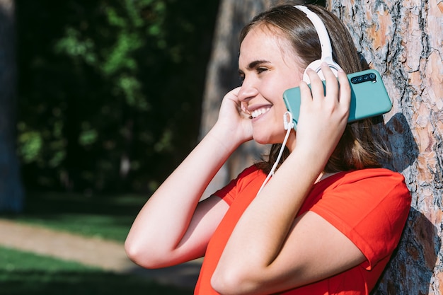 Jeune blonde portant une chemise rouge regarde le téléphone portable et écoute de la musique dans un parc