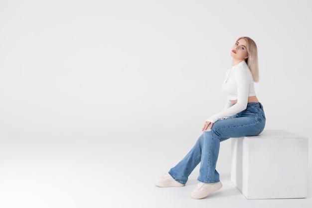 Une jeune blonde vêtue d'un pull blanc et d'un jean est assise et pose sur un fond blanc isolé en studio. Concept de mode de vie des gens. Espace de copie pour copie