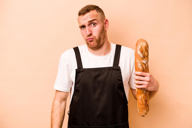 Jeune boulanger caucasien homme isolé sur fond beige hausse les épaules et ouvre les yeux confus