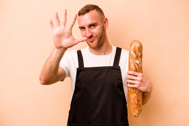 Jeune boulanger caucasien homme isolé sur fond beige souriant joyeux montrant le numéro cinq avec les doigts