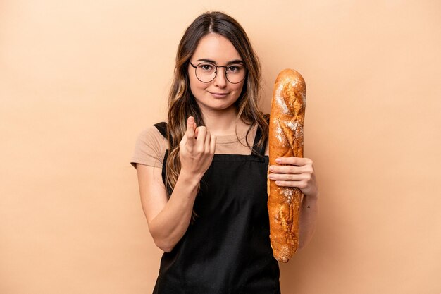 Jeune boulanger femme isolée sur fond beige pointant du doigt vers vous comme si vous invitiez à vous rapprocher