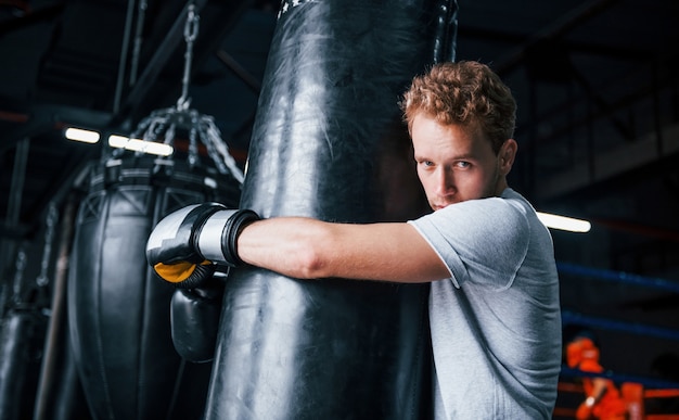 Jeune boxeur fatigué en chemise blanche et avec des gants de protection s'appuyant sur un sac de boxe dans la salle de sport.