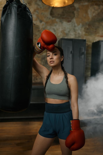 Une jeune boxeuse confiante portant des gants repose à proximité d'un sac de boxe sur l'ancienne salle de sport loft