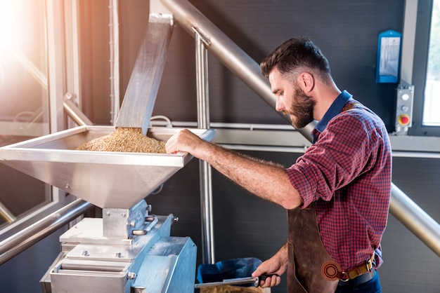 Un jeune brasseur dans un tablier en cuir contrôle le broyage des graines de malt dans un moulin d'une brasserie moderne