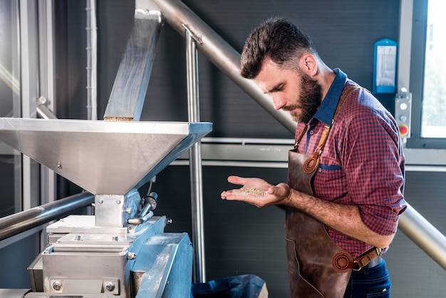 Un jeune brasseur dans un tablier en cuir contrôle le broyage des graines de malt dans un moulin d'une brasserie moderne