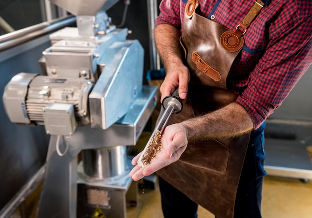 Un jeune brasseur dans un tablier en cuir contrôle le broyage des graines de malt dans un moulin d'une brasserie moderne