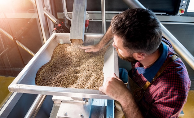 Un jeune brasseur dans un tablier en cuir contrôle le broyage des graines de malt dans un moulin d'une brasserie moderne