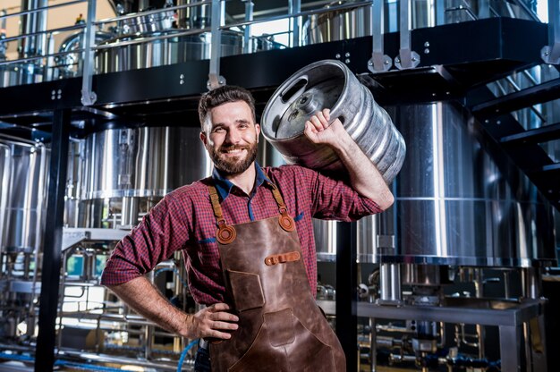 Le jeune brasseur masculin dans le tablier en cuir tient le baril avec de la bière artisanale à l'usine de brasserie moderne