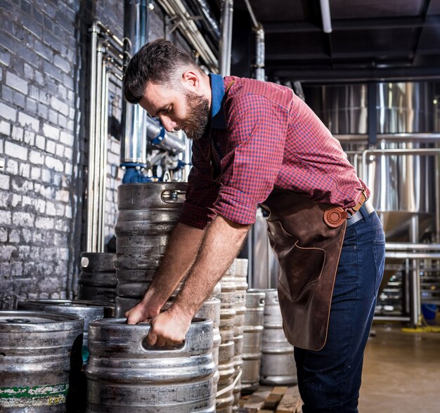 Le jeune brasseur masculin dans le tablier en cuir tient le baril avec de la bière artisanale à l'usine de brasserie moderne