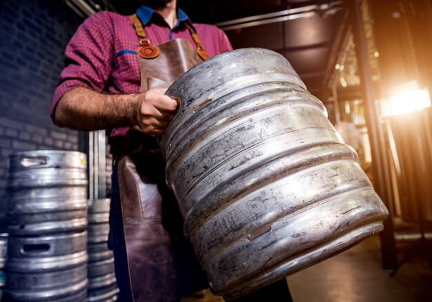 Le jeune brasseur masculin dans le tablier en cuir tient le baril avec de la bière artisanale à l'usine de brasserie moderne