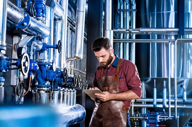 Jeune brasseur masculin en tablier de cuir supervisant le processus de fermentation de la bière dans une brasserie moderne ...