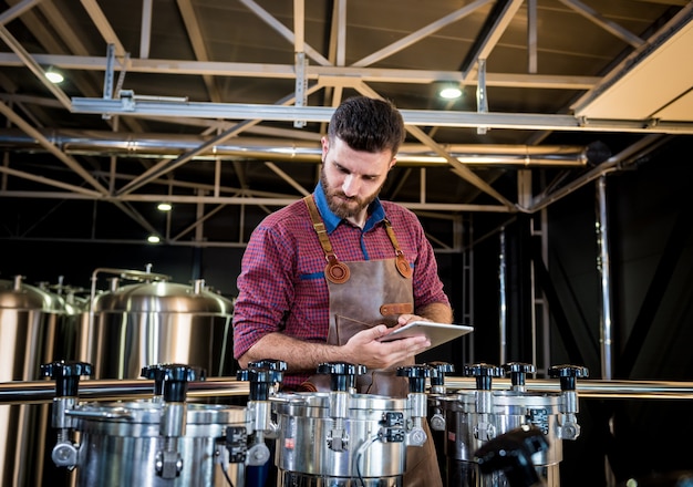 Jeune brasseur masculin en tablier de cuir supervisant le processus de fermentation de la bière dans une brasserie moderne ...