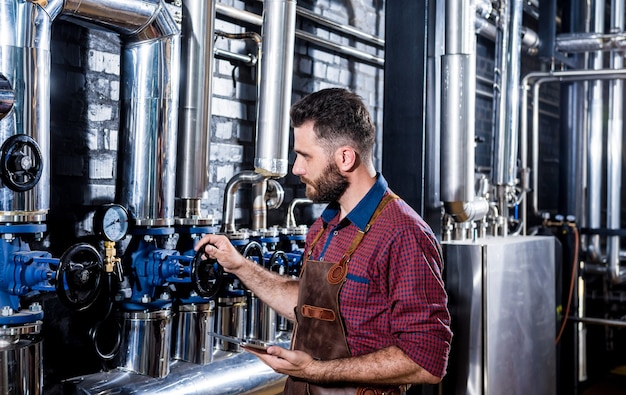 Jeune brasseur masculin en tablier de cuir supervisant le processus de fermentation de la bière dans une brasserie moderne ...