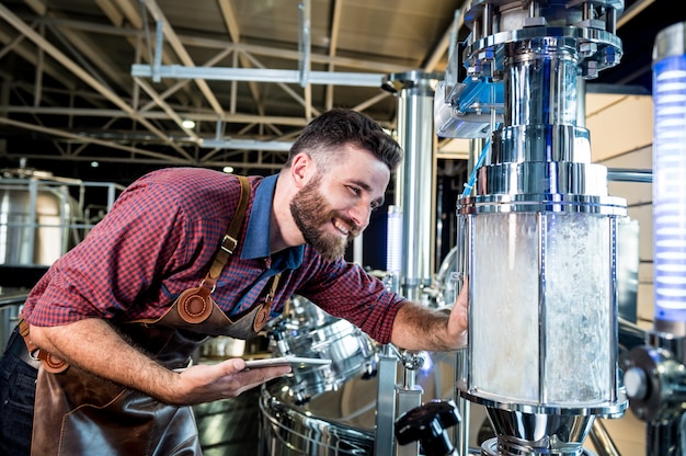 Jeune brasseur en tablier de cuir supervisant le processus de fermentation de la bière dans une brasserie moderne.