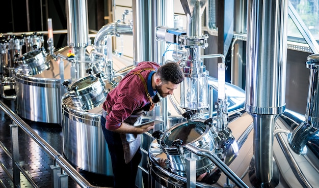 Jeune brasseur en tablier de cuir supervisant le processus de fermentation de la bière dans une brasserie moderne.