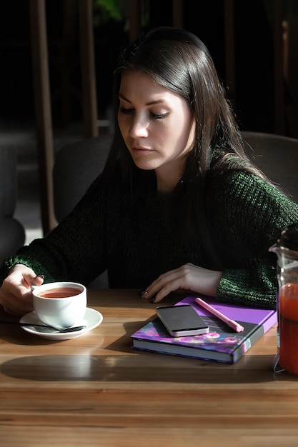 Une jeune brune d'affaires est assise à une table dans un café et boit du thé devant son téléphone