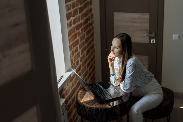 Jeune brune assise devant un ordinateur portable à la table dans la chambre