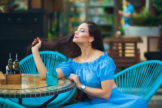 Une jeune brune aux cheveux longs dans un crop top bleu est assise pensivement à une table dans un café en plein air