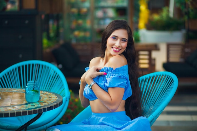 Une jeune brune aux cheveux longs dans un crop top bleu est assise pensivement à une table dans un café en plein air