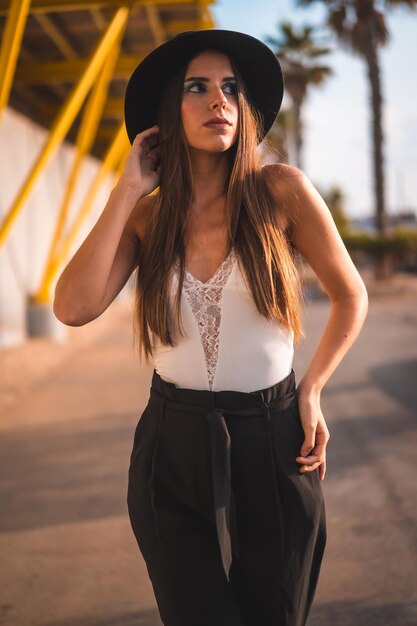 Une jeune brune dans un chapeau noir et un pantalon sur une promenade jaune