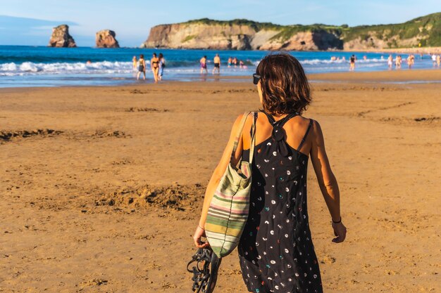 Une jeune brune en robe noire et lunettes de soleil marchant le long de la plage à Hendaye