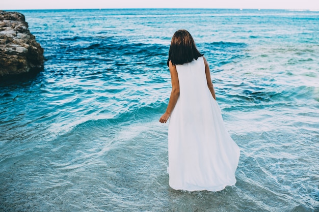 Photo jeune brune sexy dans l'eau bleue vêtue d'une robe blanche sur la plage en grèce