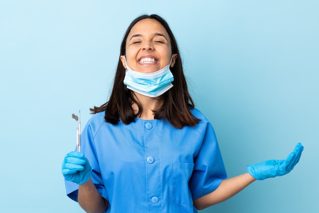 Jeune brunette dentiste métisse femme tenant des outils sur le mur isolé souriant beaucoup