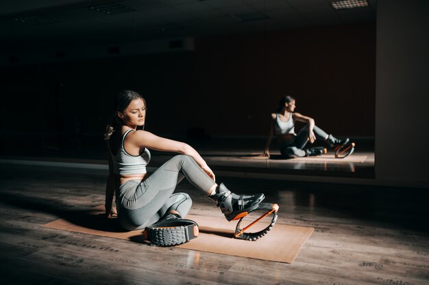 Jeune brunette fit caucasien en tenue de sport assis sur le tapis devant le miroir et portant des bottes kangoo jumps.