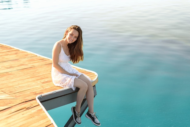 Jeune brunette souriante en robe blanche assise sur une jetée en bois contre la mer d'azur