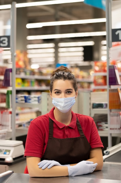 Photo jeune caissière en uniforme, masque de protection et gants assis sur le lieu de travail et attendant de nouveaux clients contre l'intérieur du supermarché