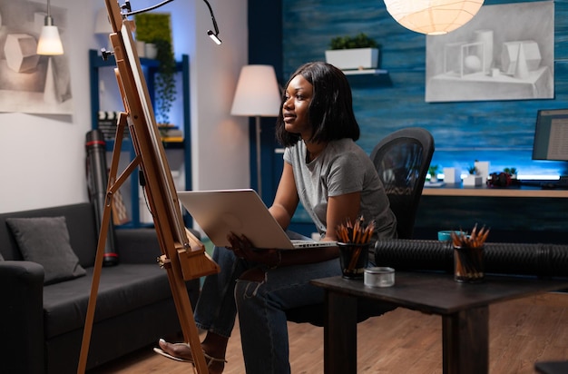 Jeune caricaturiste professionnel à la recherche d'inspiration en ligne dans un atelier à la maison avec un ordinateur portable. Artiste afro-américain examinant les notes de cours de dessin pour corriger les croquis réalisés sur un chevalet en toile.