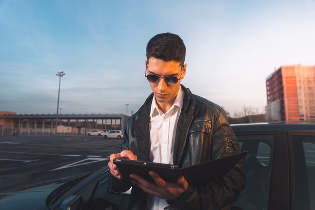 Jeune caucasien à l'aide d'une tablette numérique avec une voiture de sport derrière dans un parking.