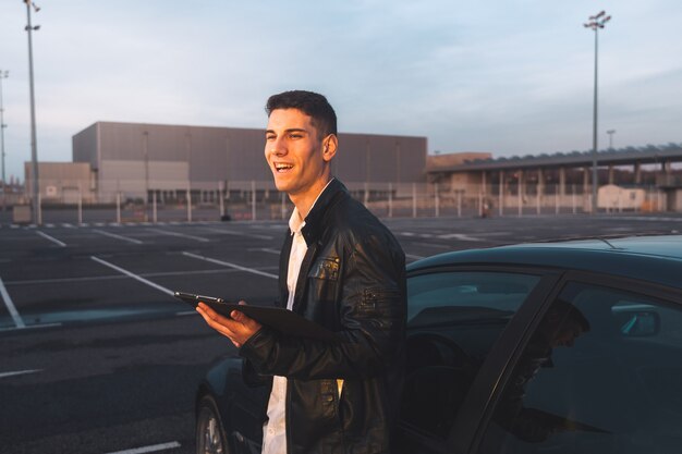 Jeune caucasien à l'aide d'une tablette numérique avec une voiture de sport derrière dans un parking.