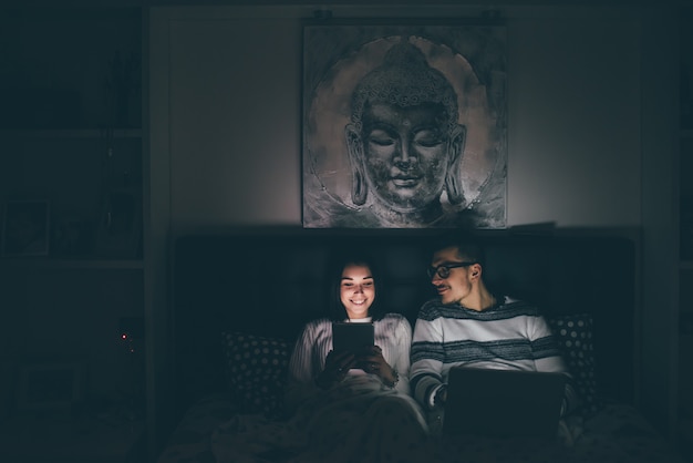 Photo jeune, caucasien, couple, coucher lit, utilisation, smartphone, et, informatique