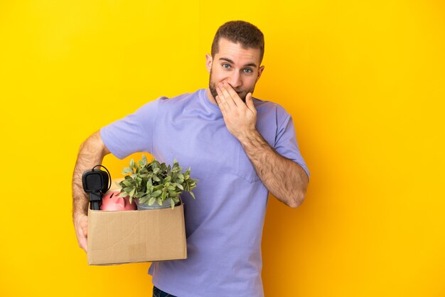 Jeune caucasien faire un mouvement tout en ramassant une boîte pleine de choses isolées sur un mur jaune heureux et souriant couvrant la bouche avec la main