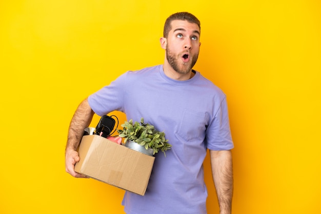 Jeune caucasien faire un mouvement tout en ramassant une boîte pleine de choses isolées sur un mur jaune en levant et avec une expression de surprise