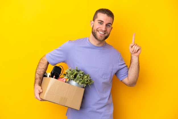 Jeune caucasien faire un mouvement tout en ramassant une boîte pleine de choses isolées sur un mur jaune pointant vers le haut une excellente idée